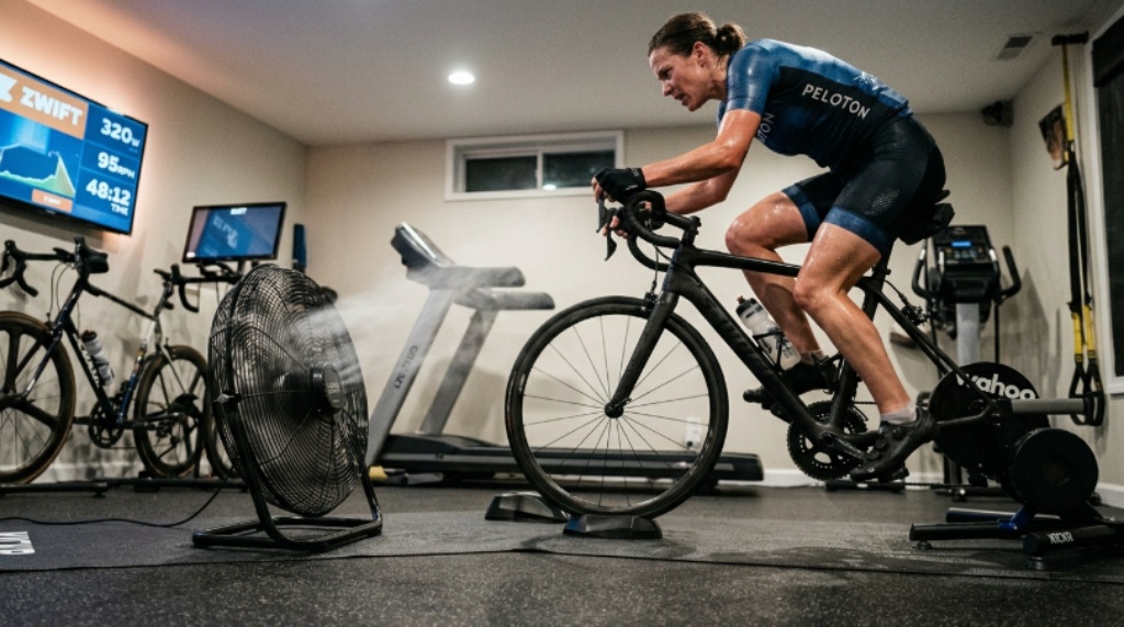 Indoor cyclist training at high intensity on a stationary bike with a large floor fan providing direct airflow in a home gym