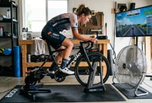 Indoor cyclist training at high intensity on a stationary bike with a large floor fan providing direct airflow in a home gym